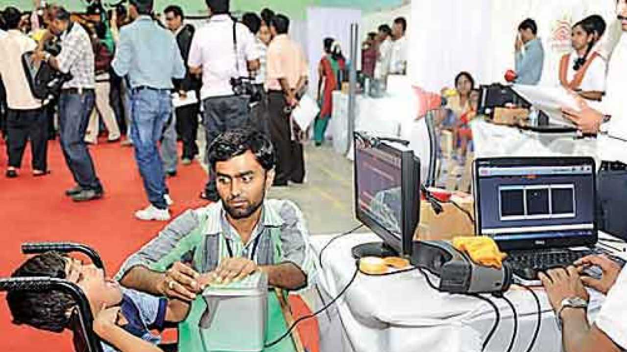 Young man presses down fingers of a boy on a wheelchair upon the biometric scanner at an AADHAAR enrolment centre